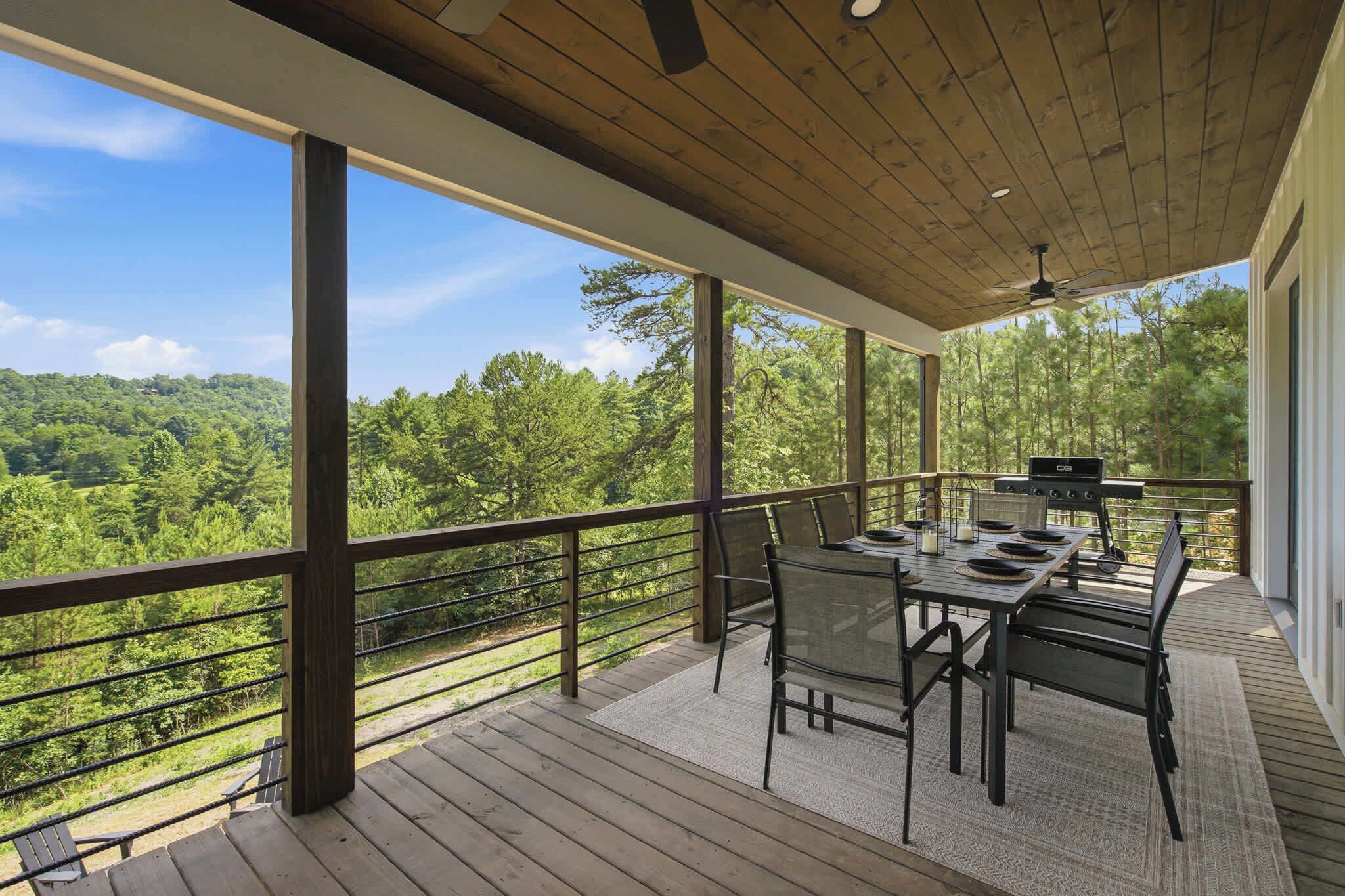 A covered outdoor deck with a dining table and chairs, a grill, and a view of a forested landscape.
