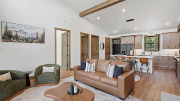 A spacious living room with a brown leather sofa, green armchairs, and a wooden coffee table. A kitchen with wooden cabinets and a dining area with bar stools is visible in the background.