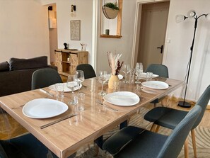 Un coin repas avec une table en bois dressée pour quatre personnes, entourée de chaises, avec un canapé et un meuble en arrière-plan.