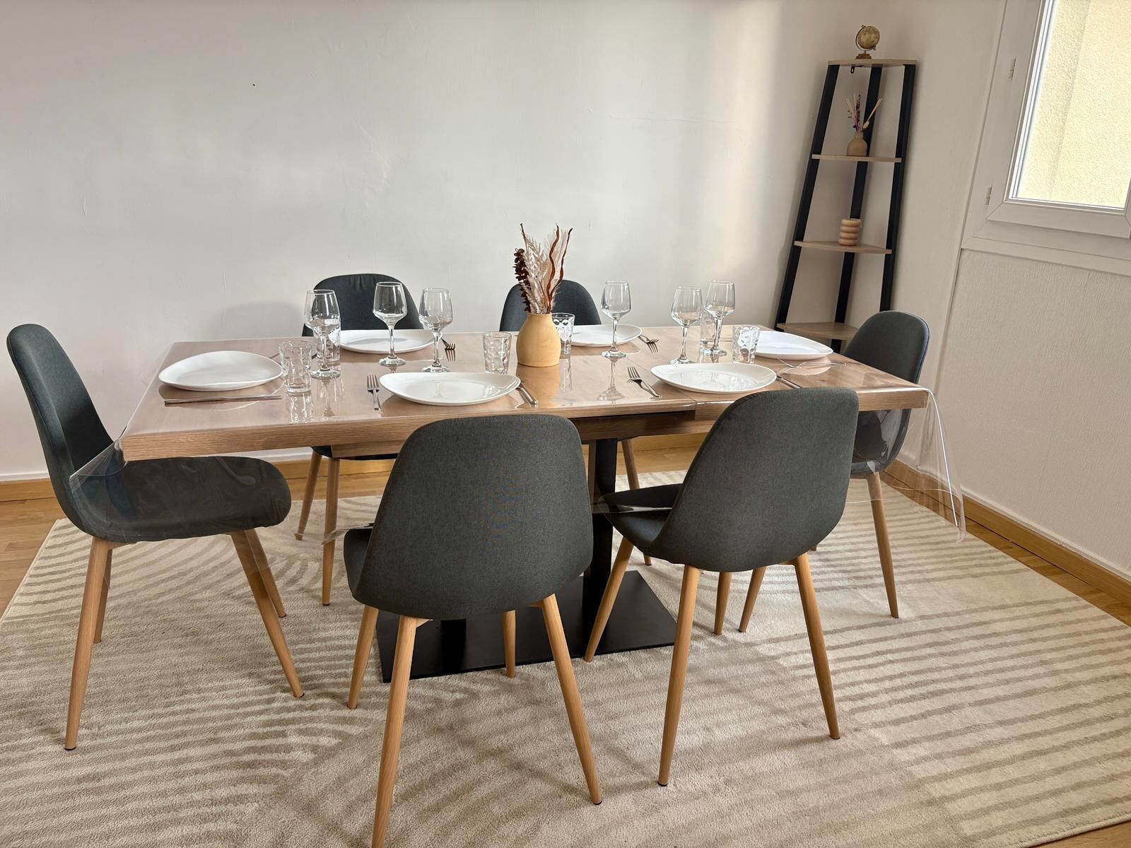 Comedor con mesa de madera preparada para cuatro personas, sillas negras y un florero con plantas secas.
