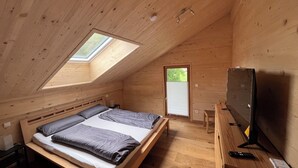 Quarto de madeira com teto inclinado, cama, escrivaninha, televisão e janela.