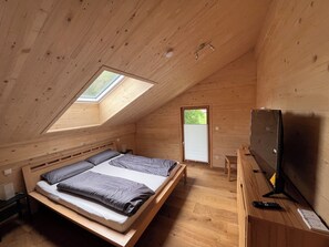 Ein Zimmer mit Holzboden und schrägen Wänden, ausgestattet mit einem Bett, einem Schreibtisch, einem Fernseher und einem Fenster.