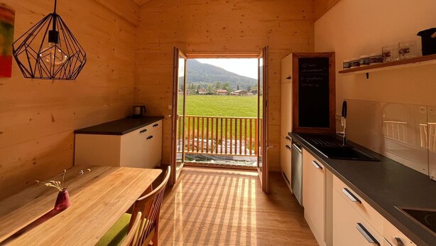 Interior de madeira com área de jantar, cozinha e varanda com vista para uma paisagem verde e montanhas.