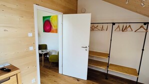Um quarto com piso de madeira, uma porta branca, uma cadeira verde, uma mesa pequena e um cabideiro com cabides.