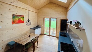 Interior de madeira com mesa de jantar, cadeiras, cozinha com fogão, lousa e vista através de uma porta.