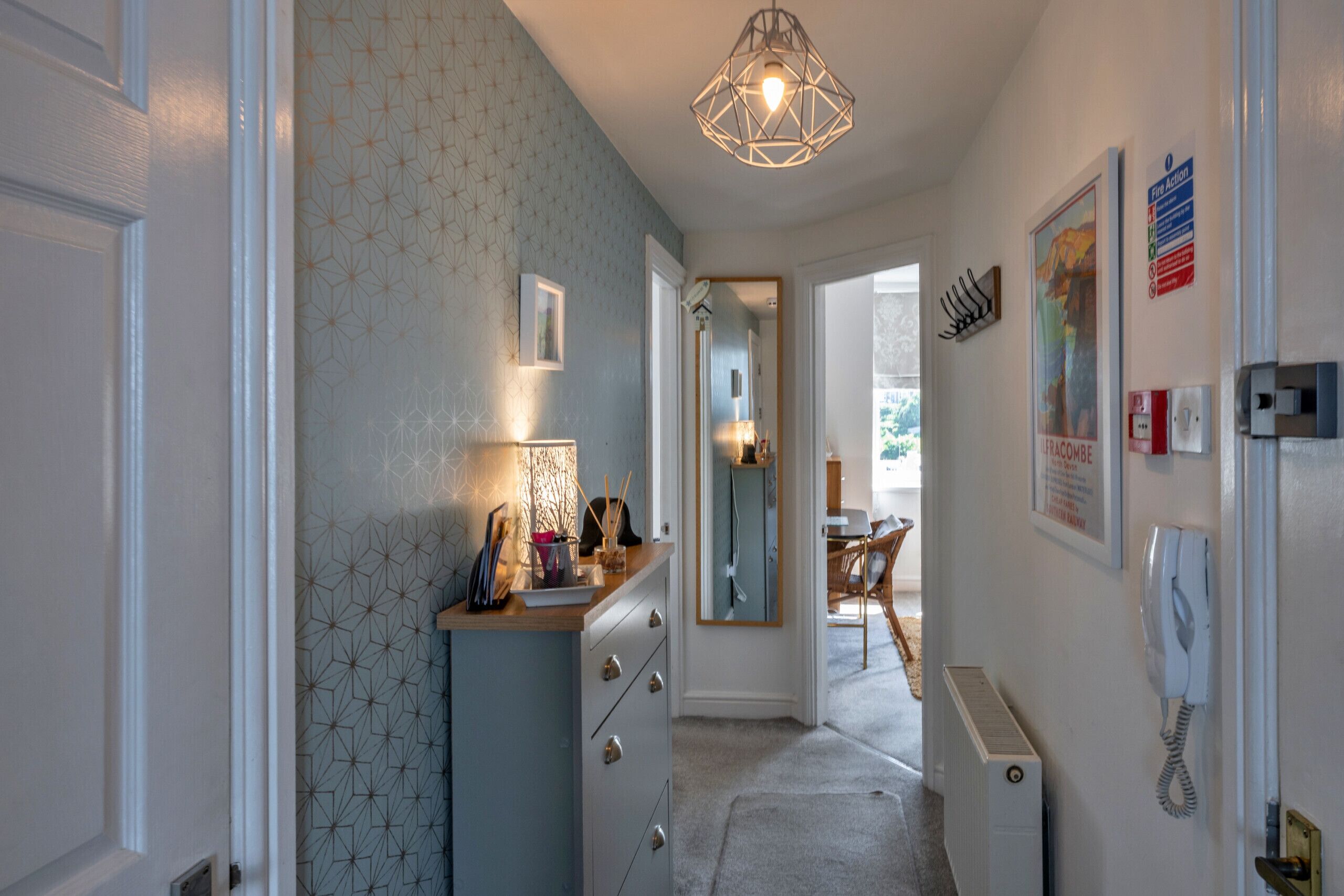 A hallway with a patterned wall, a desk with a lamp, a mirror, a door, and a telephone.