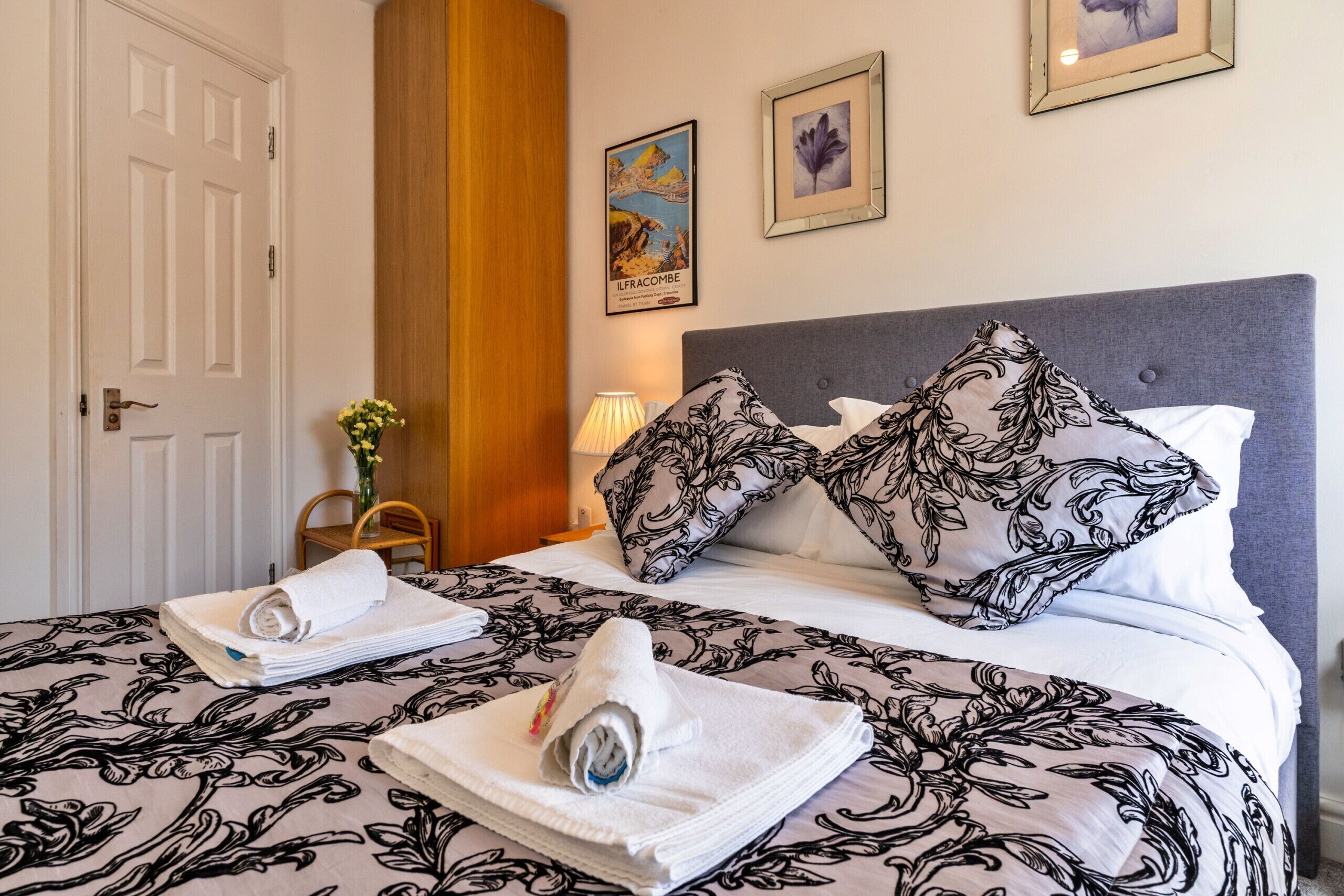 Una habitación de hotel con cama, almohadas, una silla, un florero con flores y cuadros en la pared.
