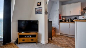 A modern living room with a flat-screen TV on a wooden stand, a kitchen area with white cabinets, and a herringbone-patterned floor. - Sea Views Apartment in Ilfracombe (Ilfracombe)