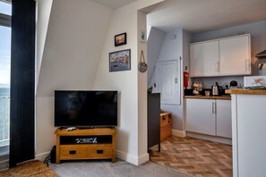 Ein modernes Wohnzimmer mit Flachbildfernseher auf einem Holzständer, einer Küchenzeile mit weißen Schränken und einem fußbodenbündigen, Zickzack-Muster-Boden.