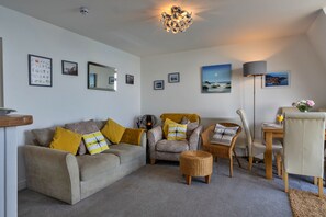 Ein Wohnzimmer mit einer Couch, Sesseln, einem Couchtisch, einem Esstisch mit Stühlen, einer Lampe und gerahmten Bildern an den Wänden.