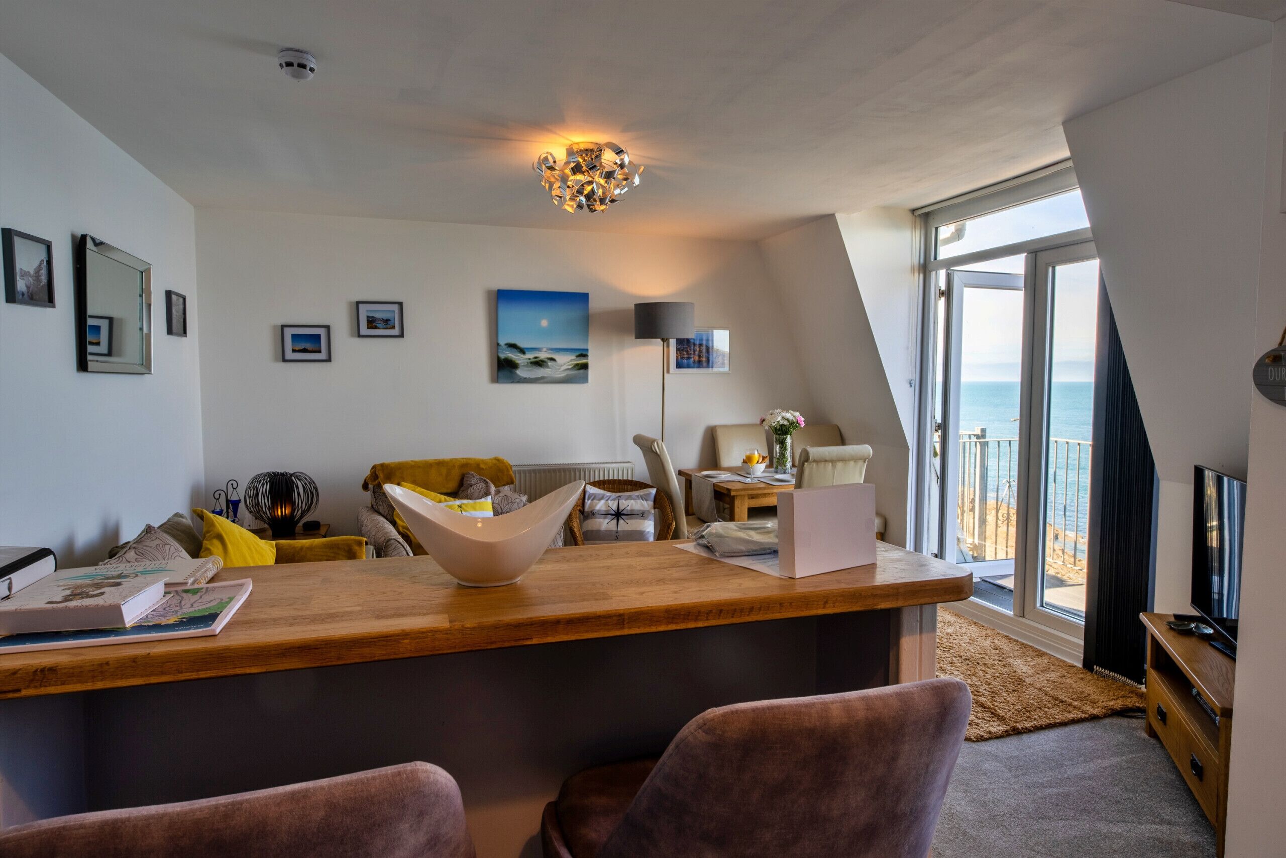 A modern living room with a wooden dining table, a sofa, and a large window offering a view of the sea.