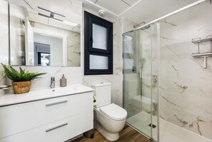 A modern bathroom with a white vanity, a toilet, a glass shower enclosure, and a marble-tiled wall. - Charming 2-bedroom apartment with AC in tranquil Villamartín (Villamartín)