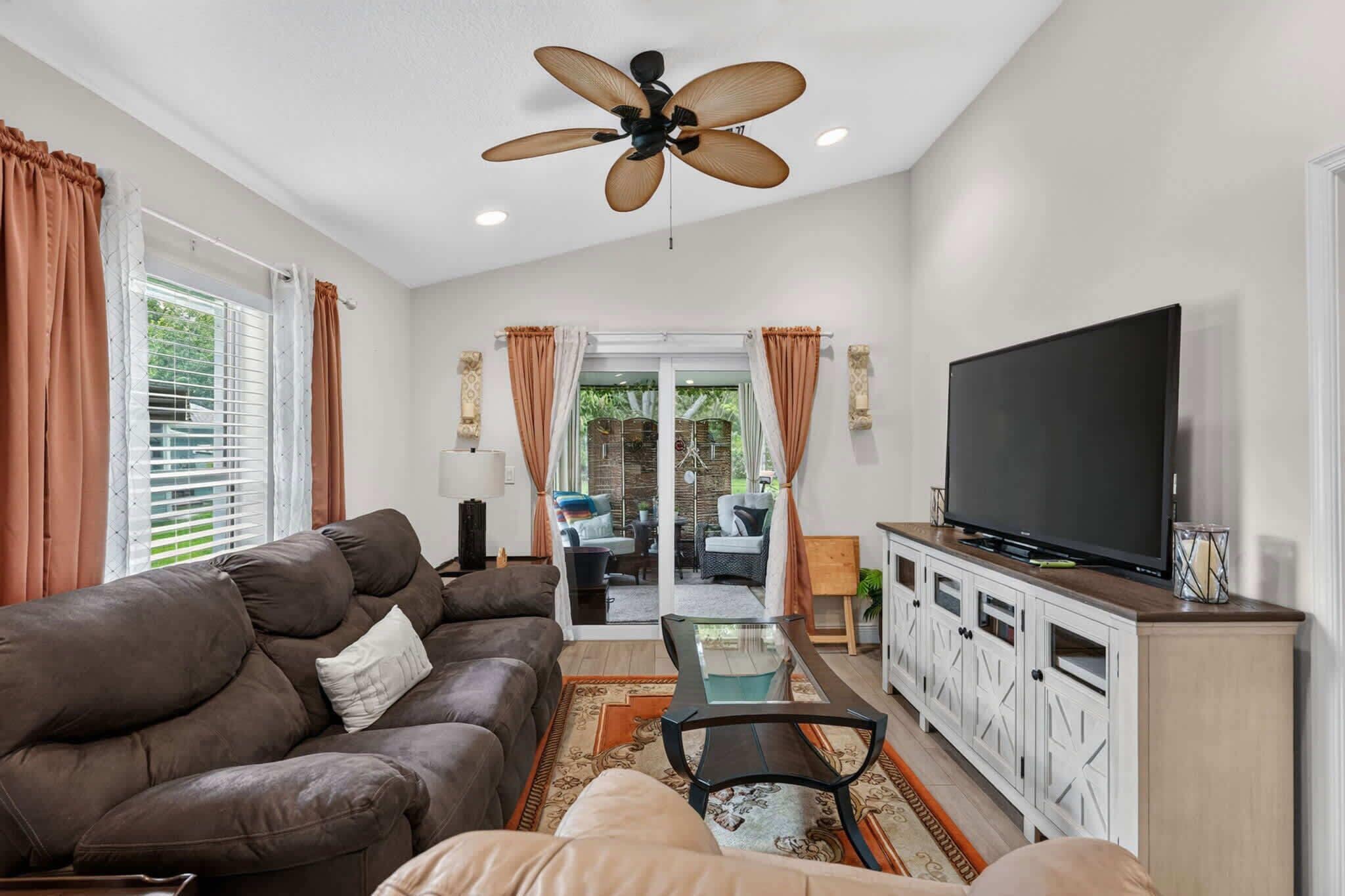 A living room with a brown sofa, a glass coffee table, a television on a cabinet, and a ceiling fan.