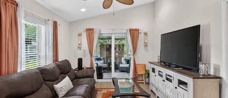 A living room with a brown sofa, a glass coffee table, a television on a cabinet, and a ceiling fan.