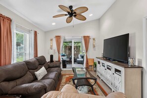 A living room with a brown sofa, a glass coffee table, a television on a cabinet, and a ceiling fan.