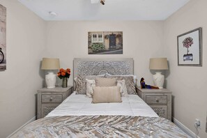 A bedroom with a bed, two bedside tables, a headboard, a wall-mounted picture, and a wall socket.