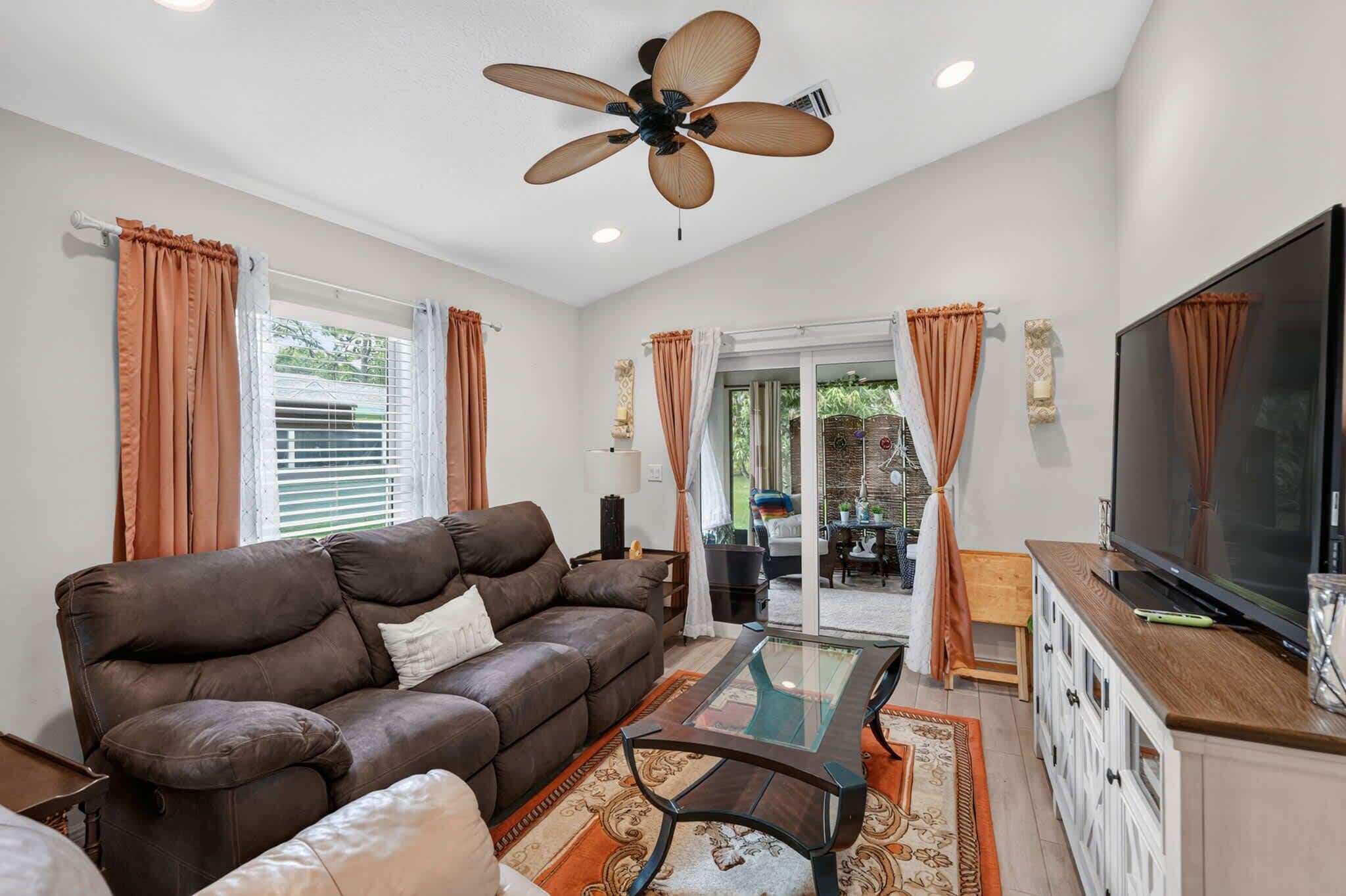 A living room with a brown sofa, a glass coffee table, a ceiling fan, and a television.