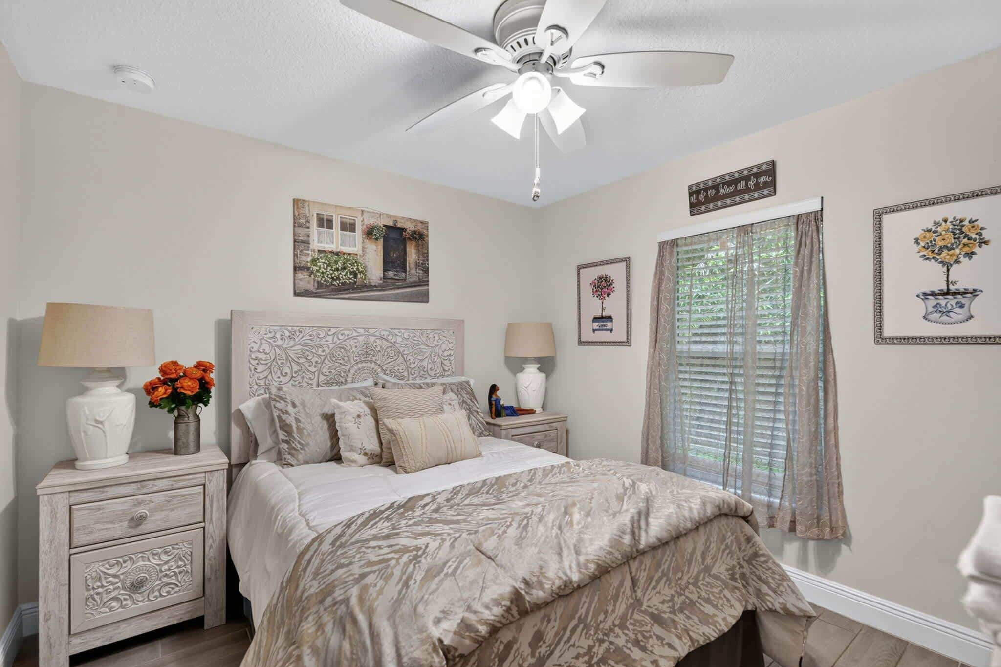 A bedroom with a bed, nightstands, a ceiling fan, a window with blinds, and wall decorations.