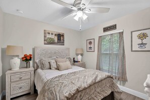 A bedroom with a bed, nightstands, a ceiling fan, a window with blinds, and wall decorations.