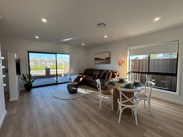 A spacious living area with a sliding glass door leading to a patio, a brown leather sofa, a dining table with white chairs, and a large window with a view of a residential area. - Summons Twenty - Sleeps 8 (Wagga Wagga)