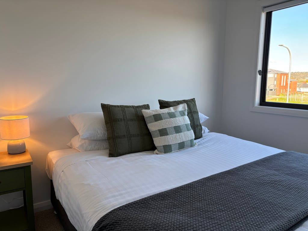 Una cama bien hecha con ropa blanca y una manta gris, una lámpara de noche y una ventana con vistas a edificios.