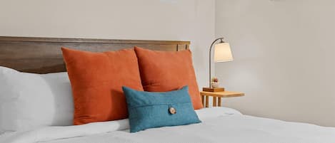 Una cama bien hecha con ropa blanca y dos almohadas naranjas, una mesita de noche de madera con una lámpara, y una pared con un cuadro enmarcado.