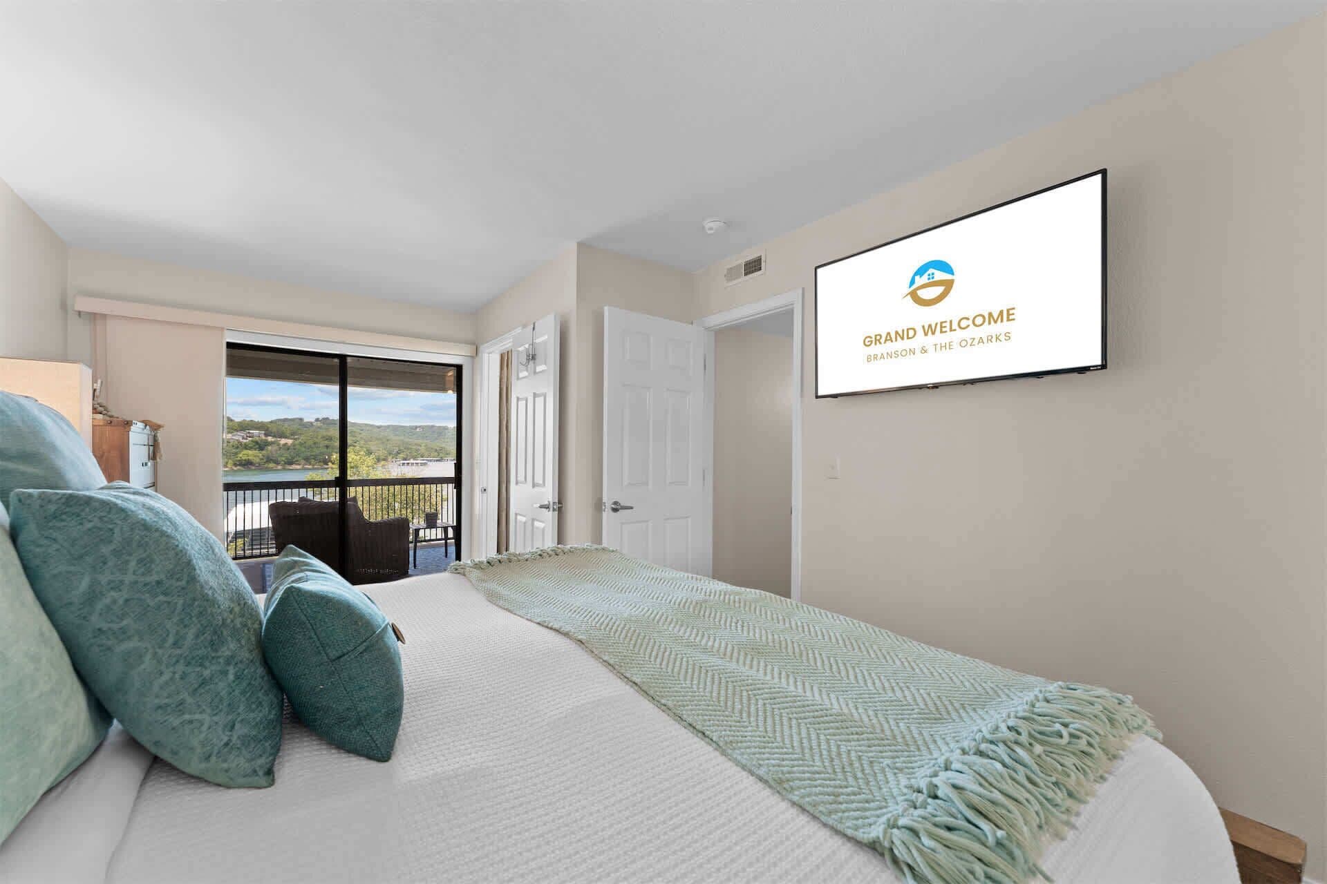 A bedroom with a bed, pillows, a TV mounted on the wall, and a balcony with a view.