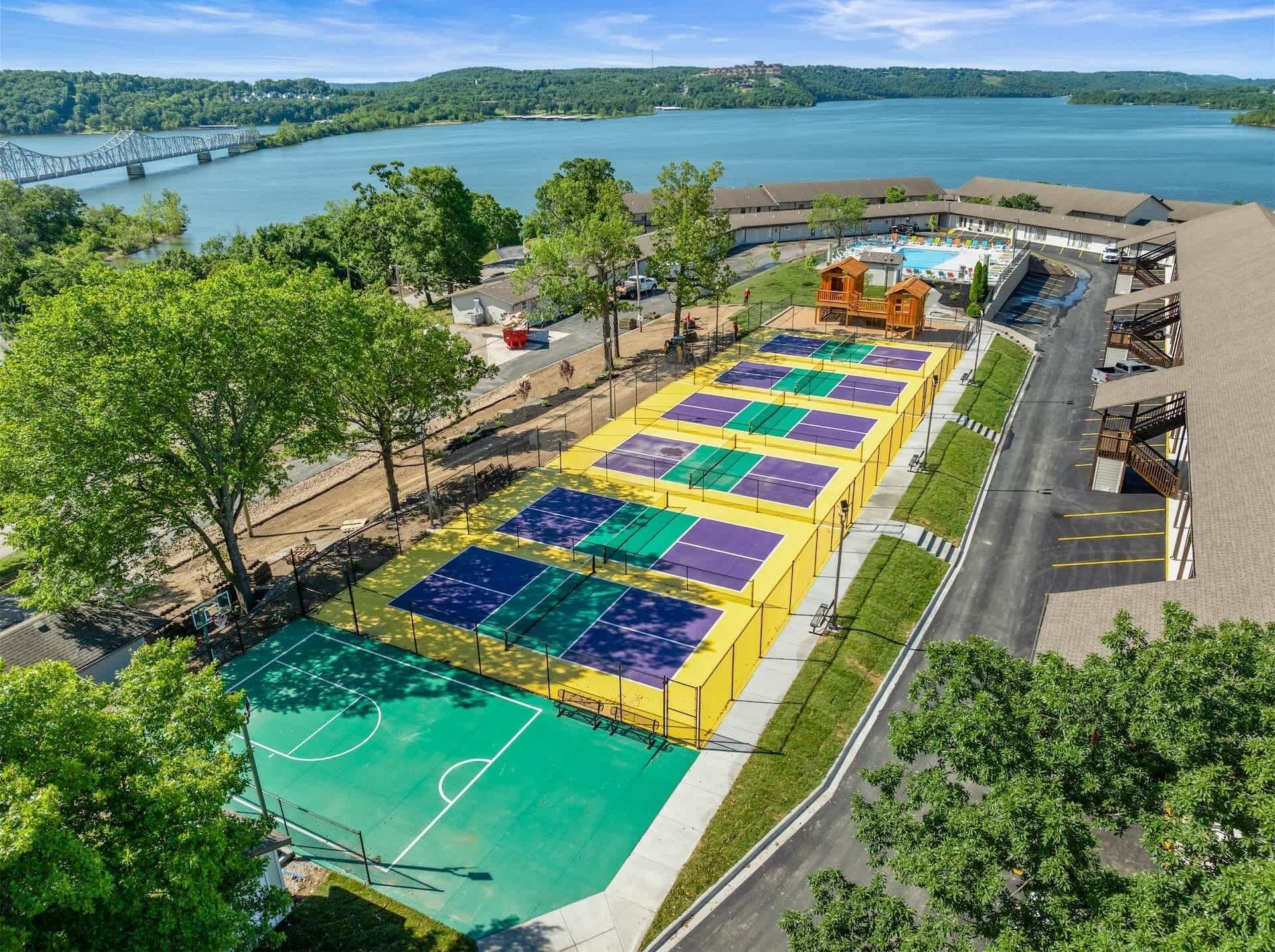 A multi-sport court complex with a basketball court, tennis courts, and a swimming pool, surrounded by trees and a view of a lake.