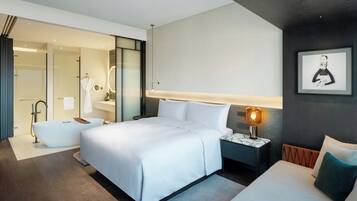 A modern hotel room with a large bed, a separate sitting area, a bathtub, and a bathroom with a glass shower enclosure.