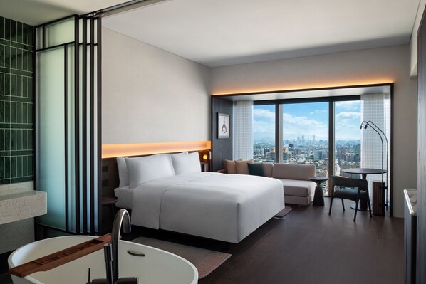 A modern hotel room with a large bed, a sitting area, a desk, and a view of the city.