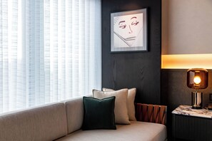 Una sala de estar moderna con un sofá, almohadones, una obra de arte enmarcada, una lámpara y una ventana con cortinas translúcidas.
