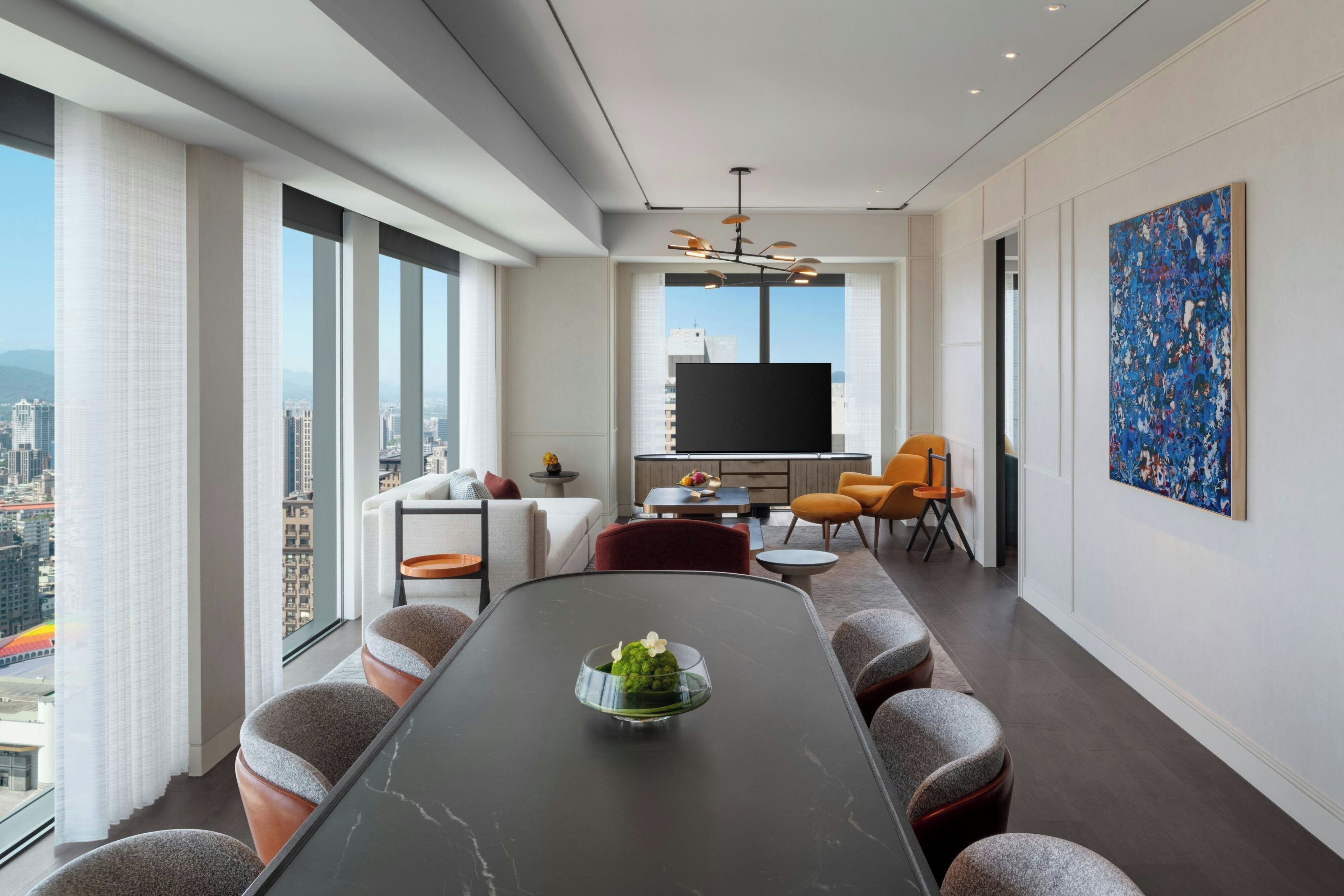 A modern living room with a large dining table, comfortable seating, a flat-screen TV, and a city view.