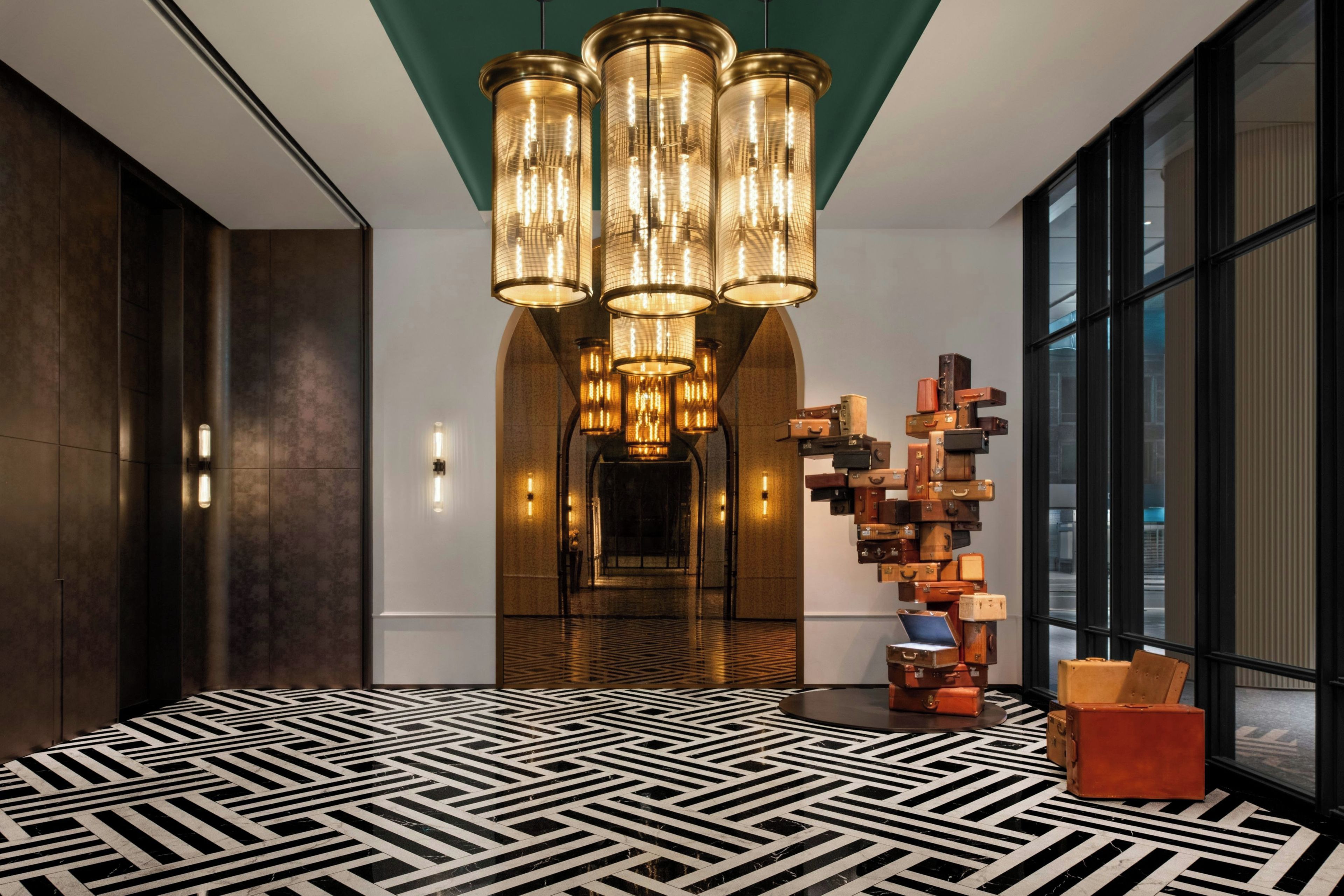 A modern lobby with a geometric patterned floor, a central chandelier, and a wall of stacked luggage.