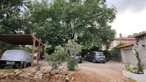 A covered outdoor area with a car parked under a shelter, surrounded by trees and a gravel path. - Villa 130 m², piscine au calme absolu – 8 pers. – Brignoles Var – DISPO AOÛT (Brignoles)