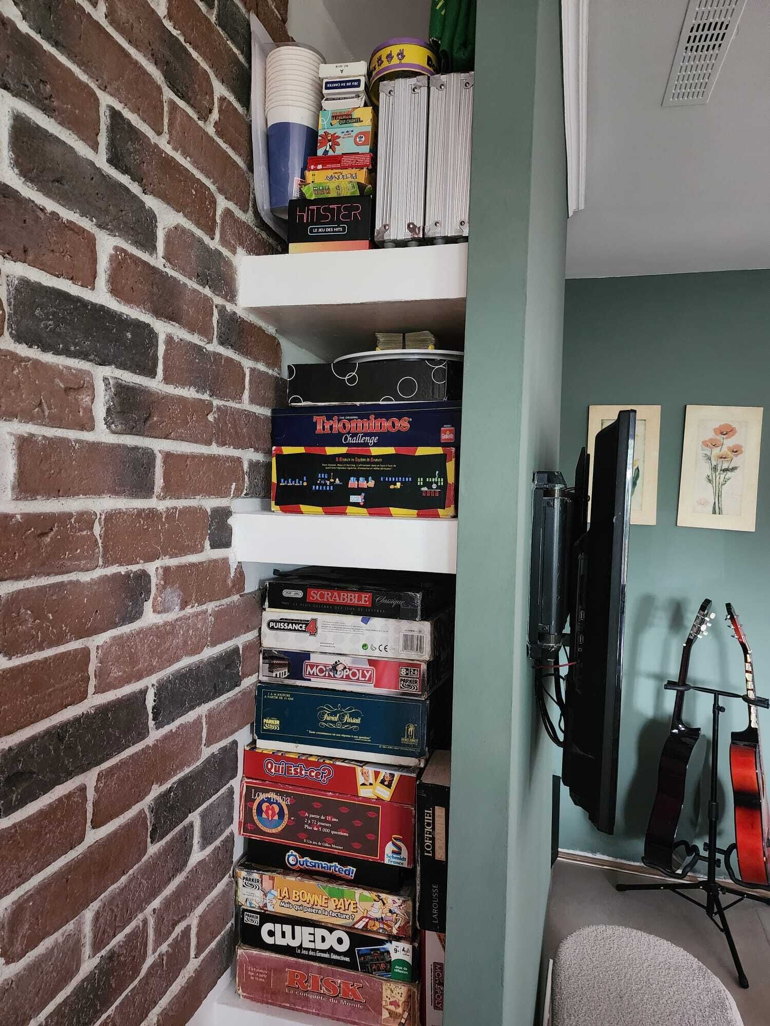 Een bakstenen muur met planken waarop spelletjes en een gitaarstandaard staan, op de achtergrond.