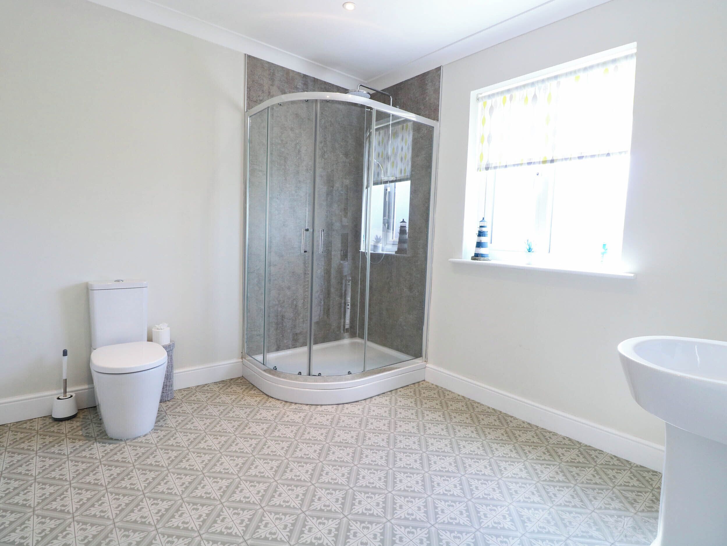 Baño con ducha de vidrio, inodoro, lavabo, baldosas de piso estampadas y ventana con cortinas.
