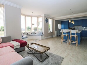Un soggiorno moderno con una zona salotto a forma di ferro di cavallo, una cucina con mobili blu e un'area pranzo con sedie in legno.