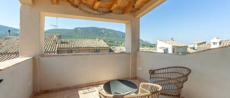 A balcony with a view of a village, a table, and two wicker chairs.