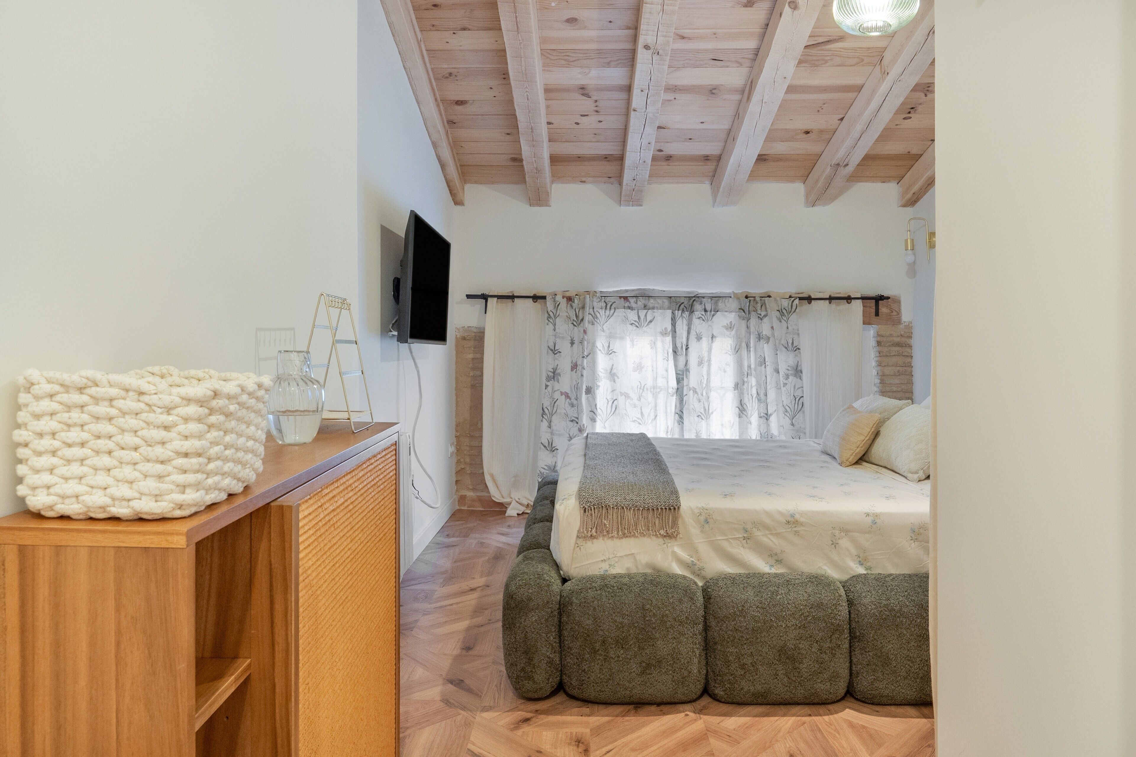Een slaapkamer met een houten plafond, een bed met een bloemenbedekking, een klein nachtkastje met een glas, en een gebreide plaid.