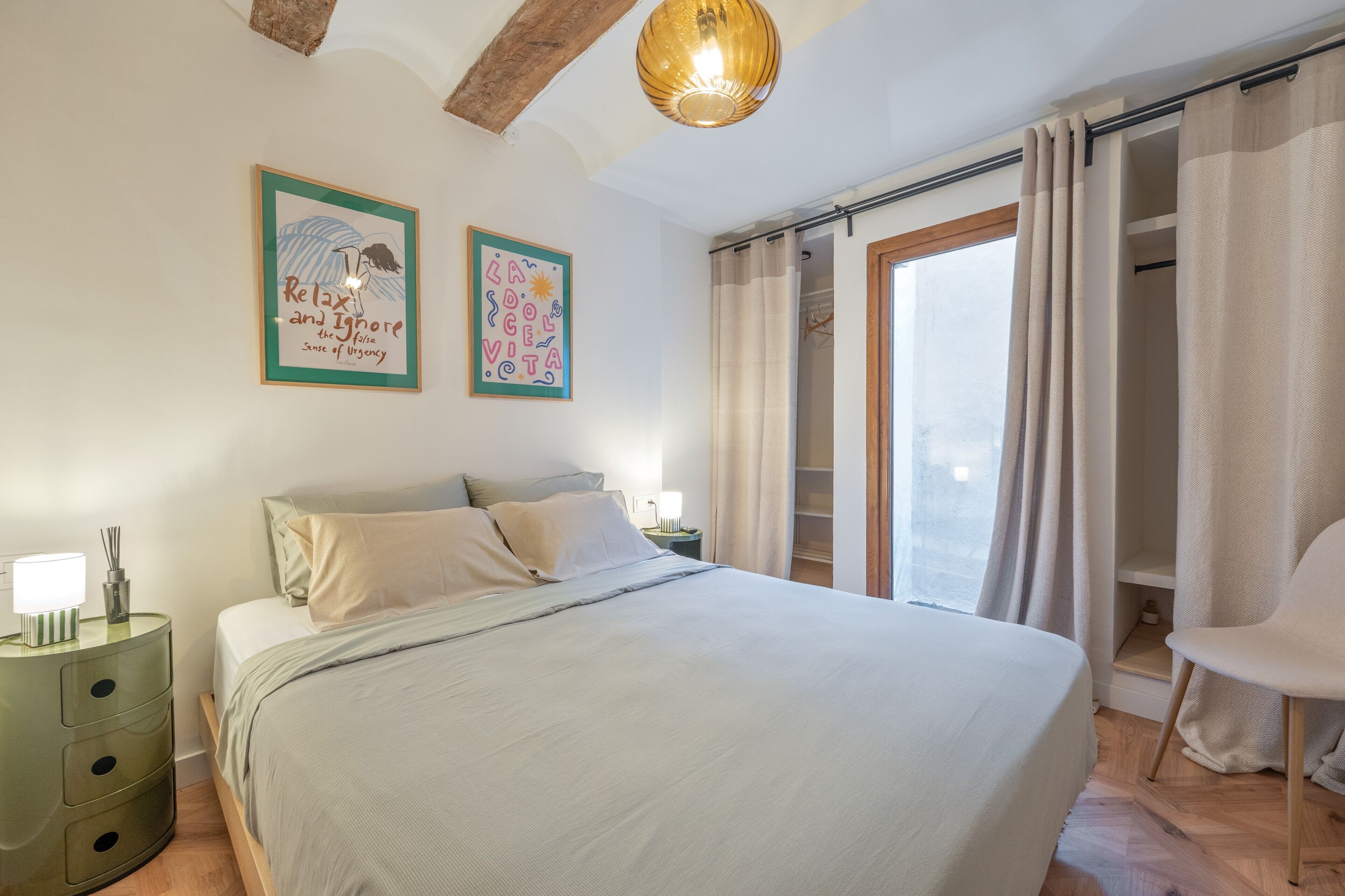 A bedroom with a bed, a nightstand, a chair, a window with curtains, and two wall art pieces.