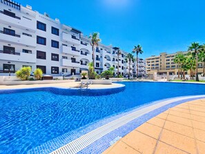 A swimming pool with clear blue water, surrounded by a white building with balconies and palm trees. - South-West Facing Balcony with Pool Views (Los Alcazares)