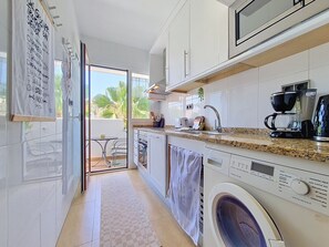 A compact kitchen with a washing machine, a coffee maker, and a small dining area with a table and chairs. - South-West Facing Balcony with Pool Views (Los Alcazares)