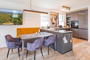 A modern kitchen with a central island, a dining area with two chairs, and a view of a forest through a window. - Modern vacation home in Zirchow for a relaxing getaway in an idyllic forest setting (Zirchow)
