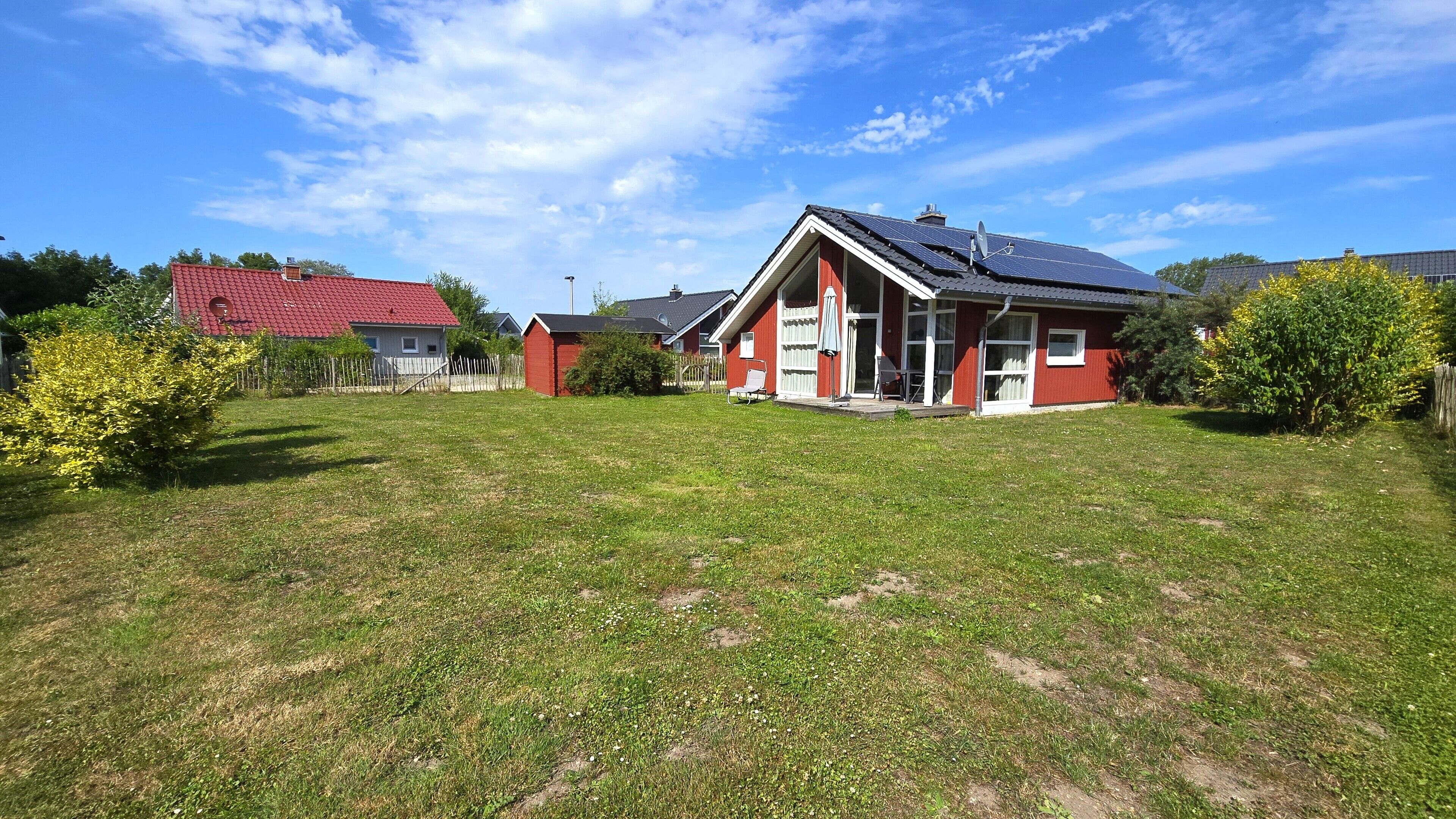 Una casa roja con techo inclinado y paneles solares, rodeada por un jardín de césped y una valla de madera.