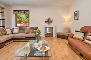 A modern living room with a sectional sofa, a glass coffee table with magazines, a fireplace, and a view of greenery outside.