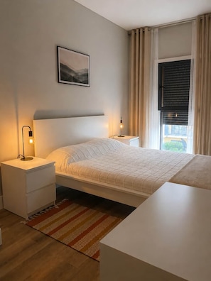 A bedroom with a bed, bedside tables, a nightstand, a window with curtains, and a wall-mounted picture.