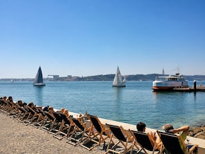A waterfront with lounge chairs along the edge, sailboats on the water, and a ferry docked at the pier.