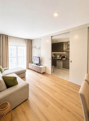 A modern living room with a sofa, a television, and a kitchen area visible through an open door.