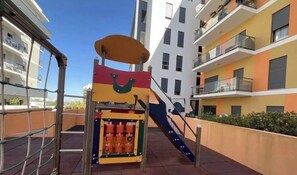 Un terrain de jeux équipé d’un toboggan et de structures d’escalade, entouré d’immeubles résidentiels à plusieurs étages.