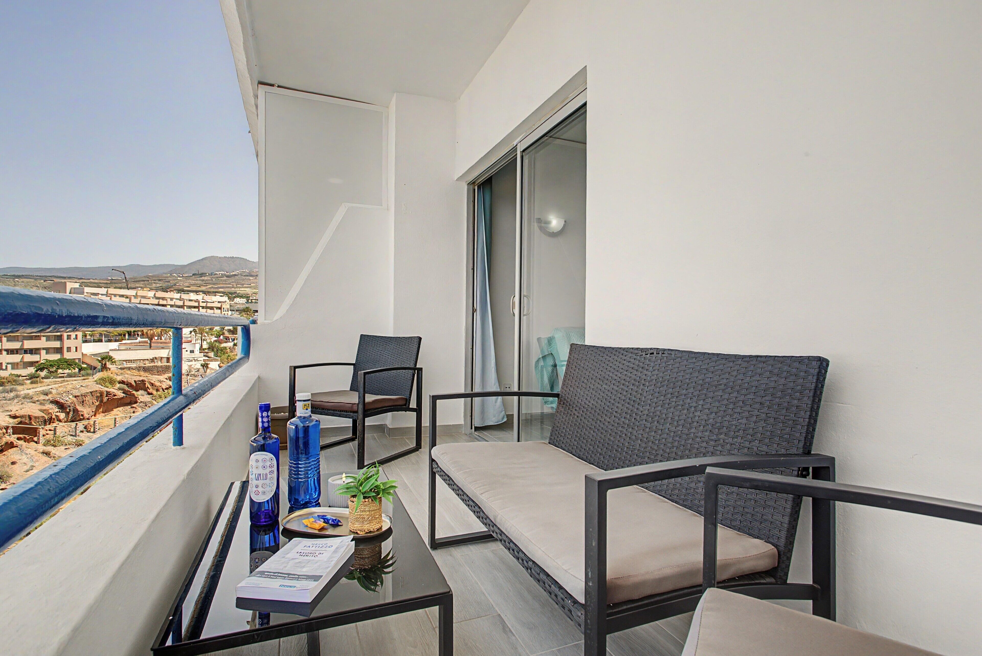Un balcón con sofá, sillas, una mesa con bebidas y un menú, y vistas de un paisaje costero.